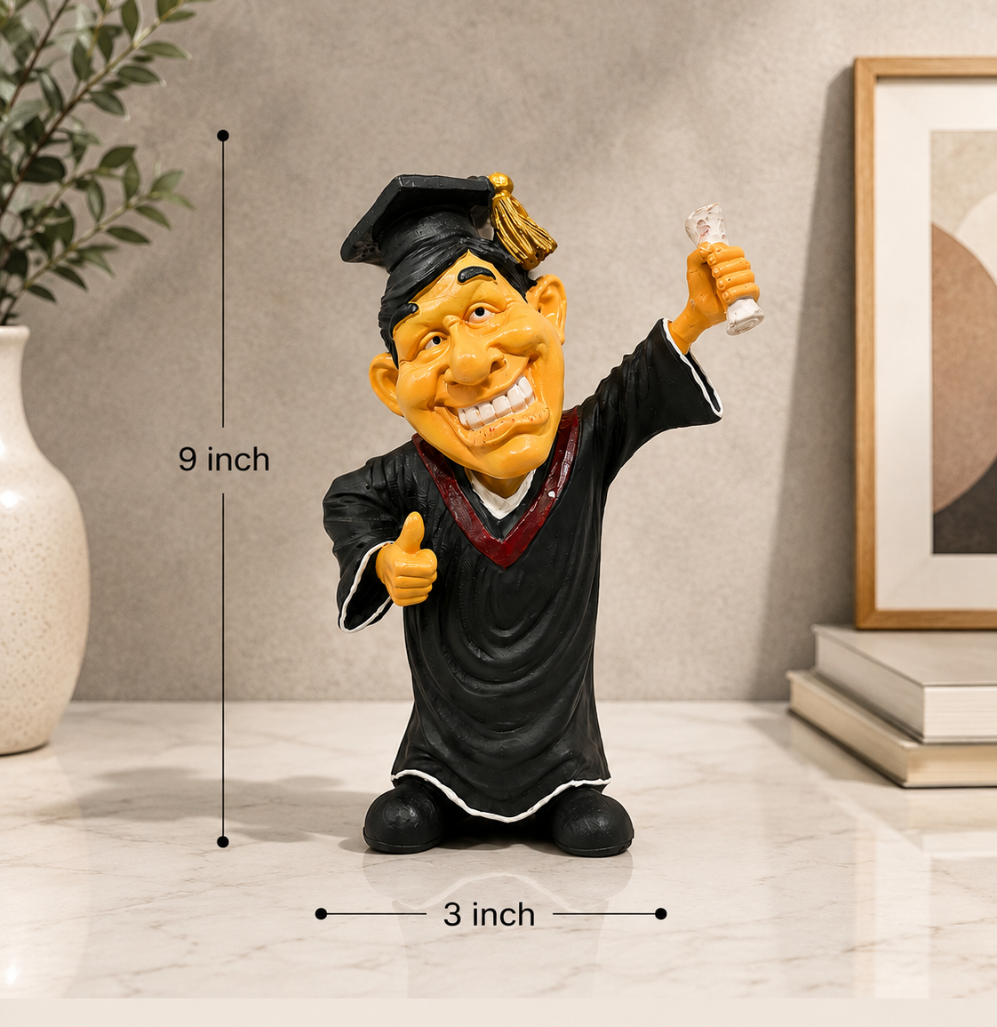 A personalized graduation figurine of a cartoon graduate in a black gown and cap holding a diploma, shown on a marble surface with dimension markers indicating 9 inch height and 3 inch width. The background features a neutral wall, a plant, a vase, and books.