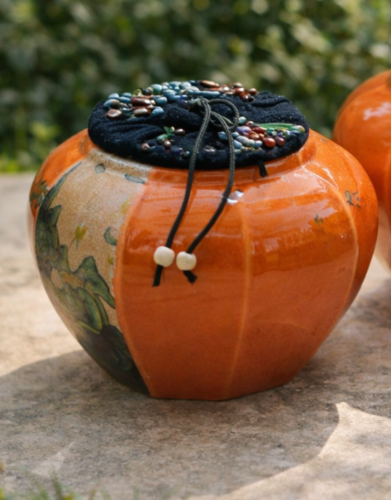 Two decorative orange pots with black lids on a stone surface with greenery in the background.