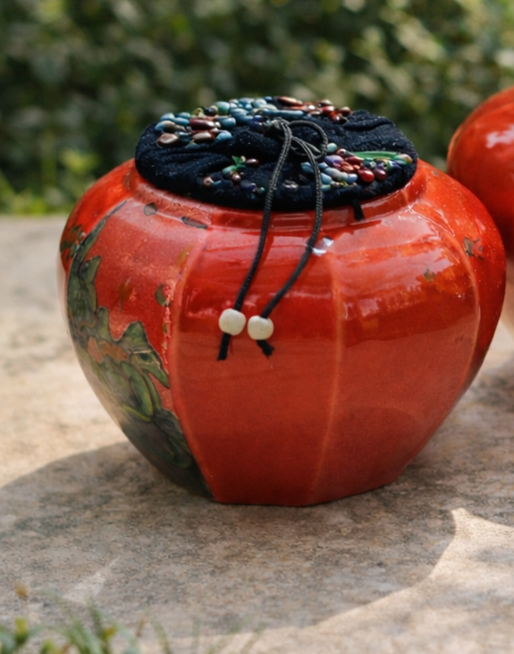Two red ceramic pots with decorative lids on a stone surface, blurred greenery in the background.