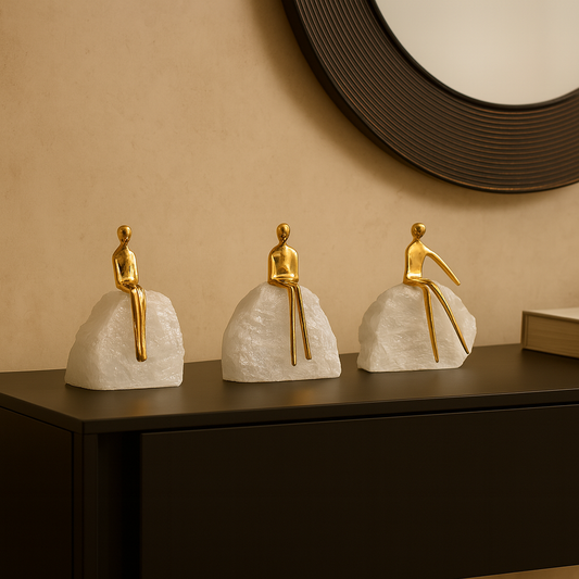 Three gold figurines on white rock-like bases on a dark surface with a beige wall in the background.