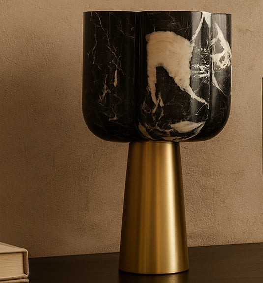 Table lamp with black marble shade and gold base on a dark surface against a beige wall.