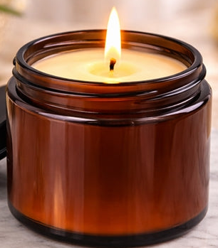 Candle in an amber jar with a black lid on a marble surface, surrounded by flowers.