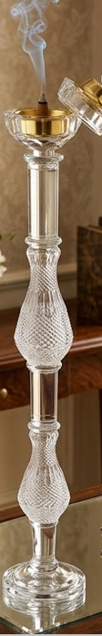Decorative glass candlestick with a small flame on a reflective surface, with a mirror and books in the background.