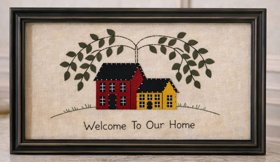 Framed cross-stitched artwork with houses and 'Welcome To Our Home' text on a marble surface.